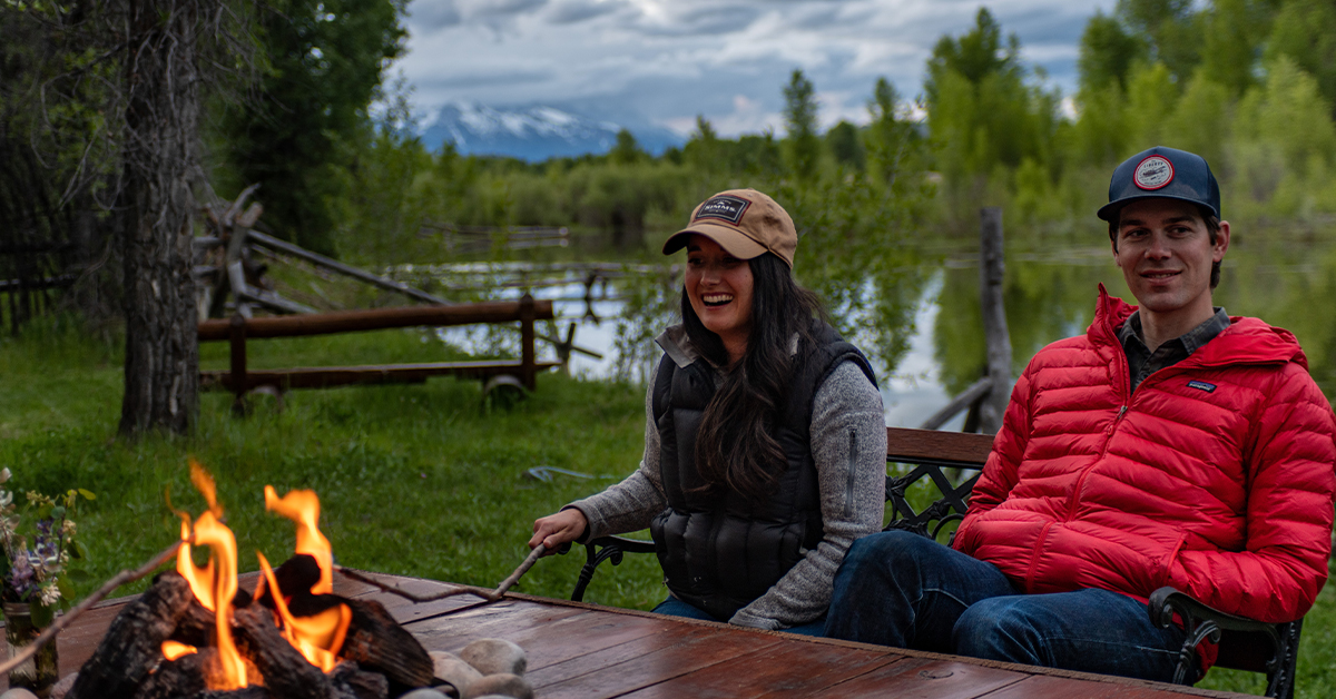 A Young Couple Sits On A Bench Along The Snake River While Enjoying A Campfire Against The Backdrop Of The Mountains