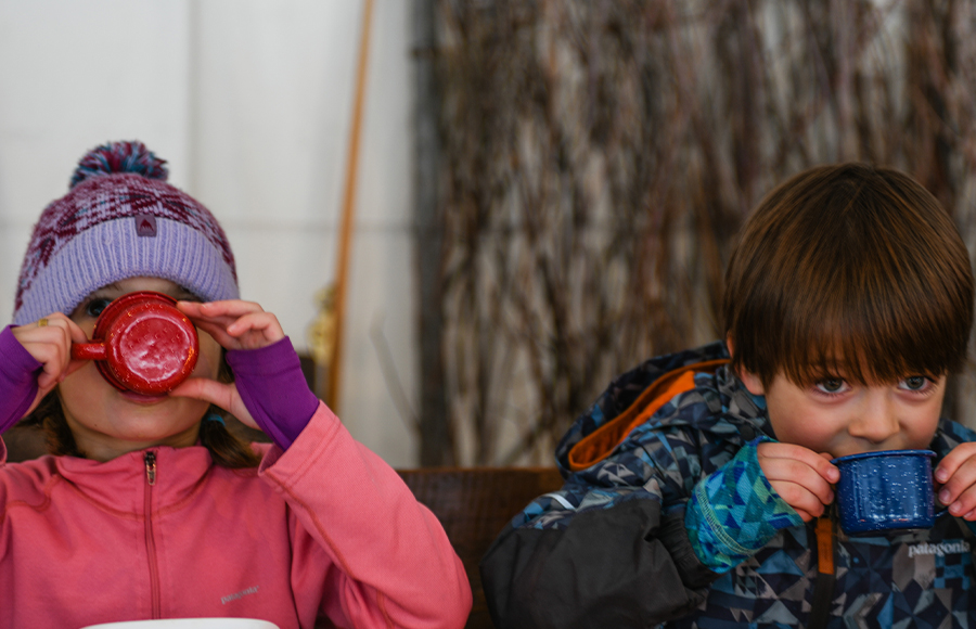 Two Children Dressed In Winter Snowsuits Sip On Cups Of Hot Cocoa
