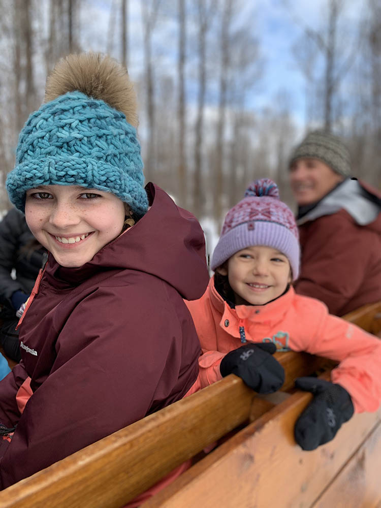 Two Girls Dressed In Winter Clothing Enjoy A Wooden Sleigh Ride In Jackson Hole, Wyoming